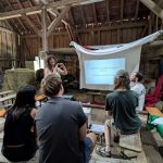 Liz Pendleton holds a workshop on responses to media and AI enshittification. She makes a heart shape with her hands, and stands in front of a screen with a projected slideshow. The slide reads ‘Self governed platforms beyond BigTech. Fediverse introduction.’. 4 people are sat on wooden benches watching Liz. They are in a barn, there is a haybale to the left of Liz.