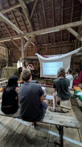 Liz Pendleton holds a workshop on responses to media and AI enshittification. She makes a heart shape with her hands, and stands in front of a screen with a projected slideshow. The slide reads ‘Self governed platforms beyond BigTech. Fediverse introduction.’. 4 people are sat on wooden benches watching Liz. They are in a barn, there is a haybale to the left of Liz.